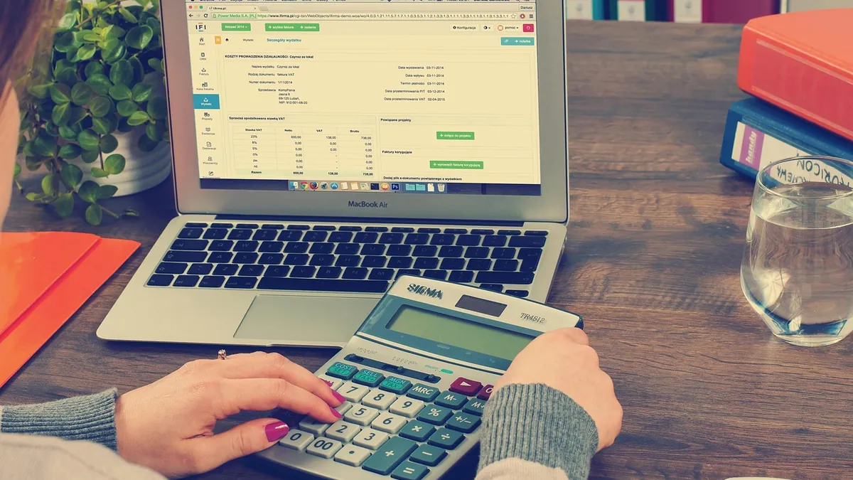 An accountant checks figures on a desk calculator beside a spreadsheet open on her laptop