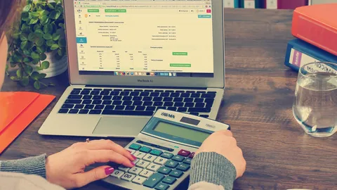 An accountant checks figures on a desk calculator beside a spreadsheet open on her laptop