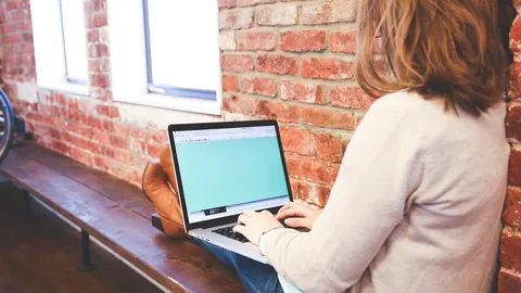 Woman typing a message on a laptop by a brick wall, working on her own schedule