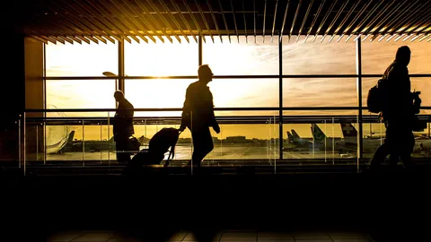 Siluetas de viajeros con maletas caminando por una terminal de aeropuerto al atardecer, rumbo al extranjero