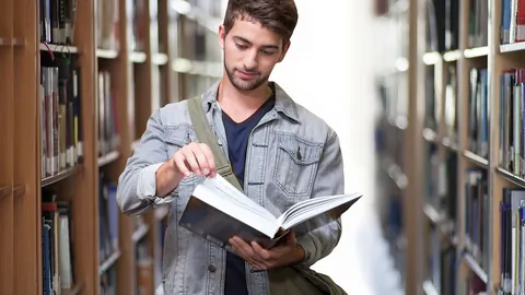 Étudiant lisant un livre entre les rayons d'une bibliothèque universitaire en préparant sa candidature en master