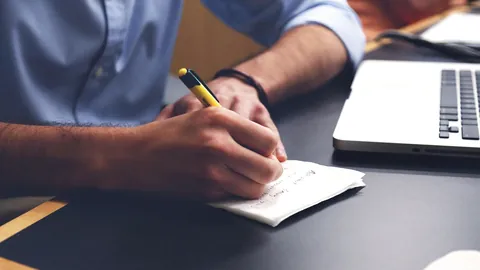 Man writing notes with a pen beside his laptop while drafting his CV step by step