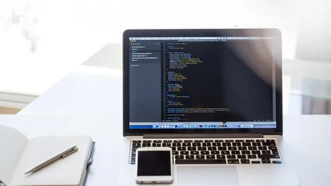 Laptop showing program code on a bright desk with a smartphone and an open notebook