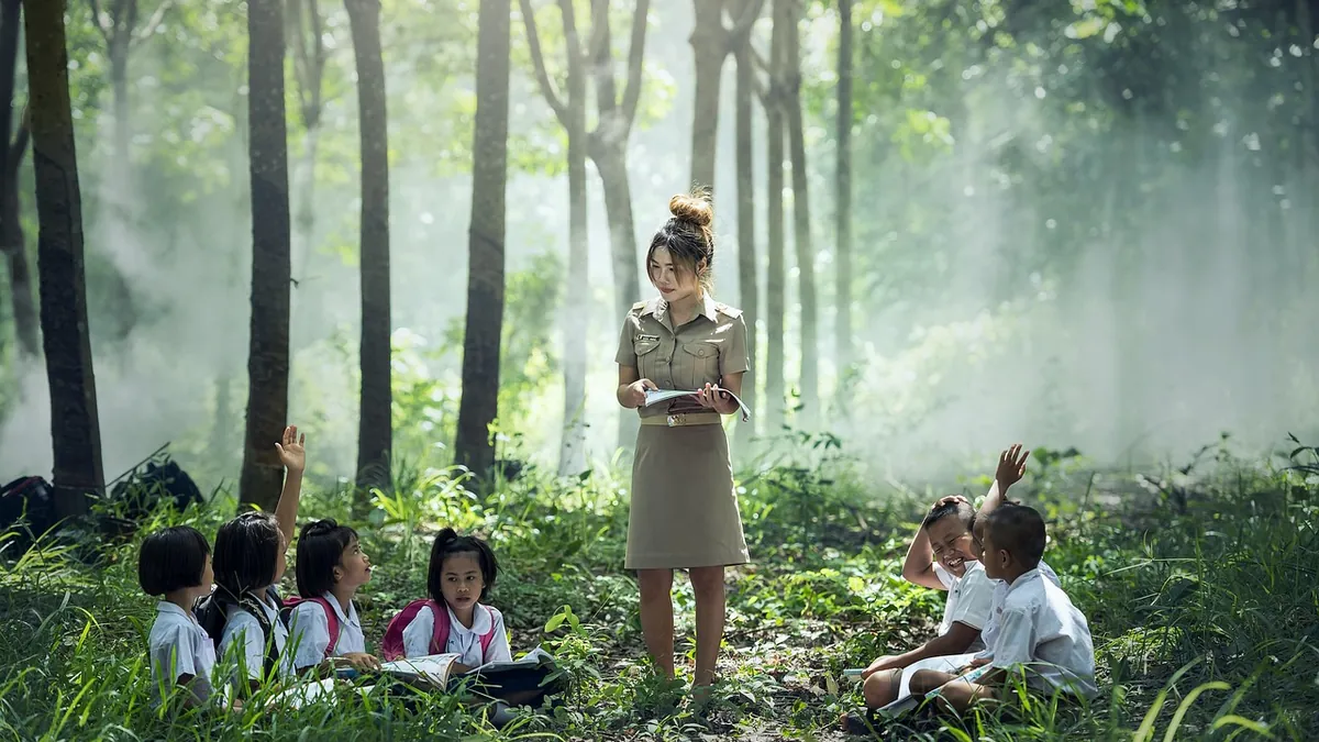 A teacher reads to a group of children sitting on the grass during an outdoor lesson among trees