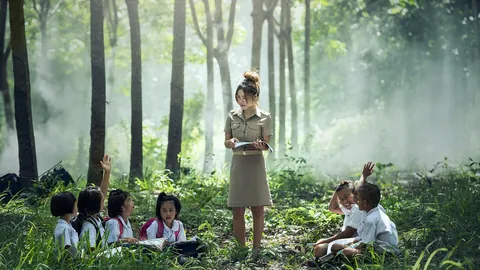 A teacher reads to a group of children sitting on the grass during an outdoor lesson among trees